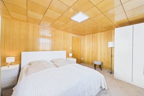 Un dormitorio con techo de madera, una cama grande, una mesita de noche con lámpara, un taburete pequeño y un armario.