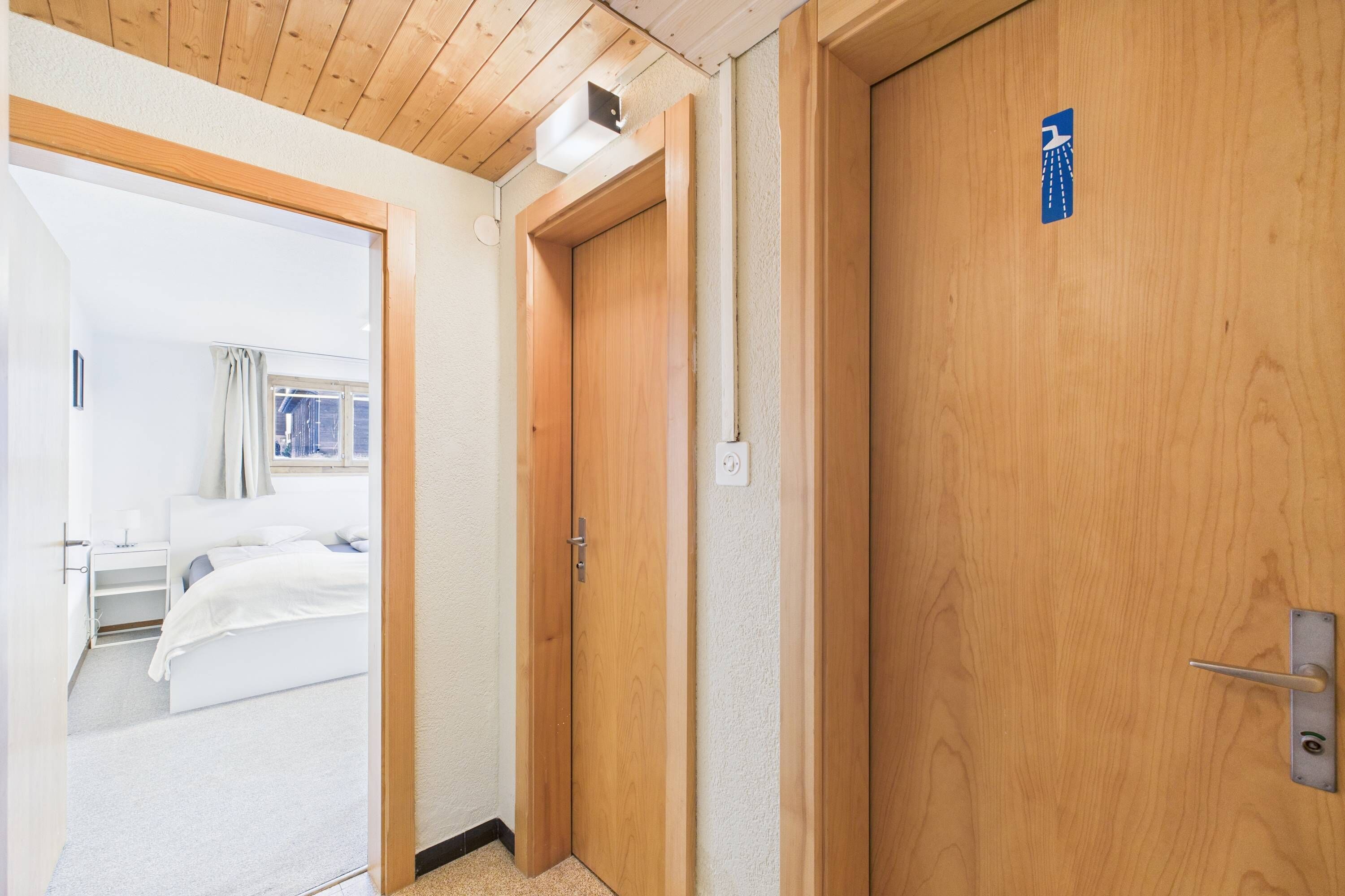 A hotel room with a wooden door, a bed, a bedside table, and a window with curtains.