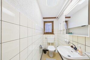 Een badkamer met witte tegels, een toilet, een wastafel en een raam met een kantonsluier.