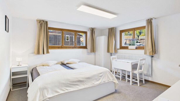 A bedroom with a bed, a desk, and two windows with curtains. - Mountain View Studio Near Saas-Fee Gondola (Saas-Grund)