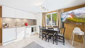 Une cuisine moderne avec des meubles blancs, une table à manger avec des chaises, une bouilloire rouge et une grande fenêtre avec des rideaux.
