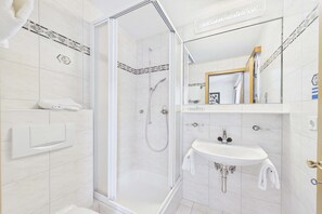 Un baño con ducha de mampara de vidrio, un lavamanos blanco y un espejo con marco de madera.
