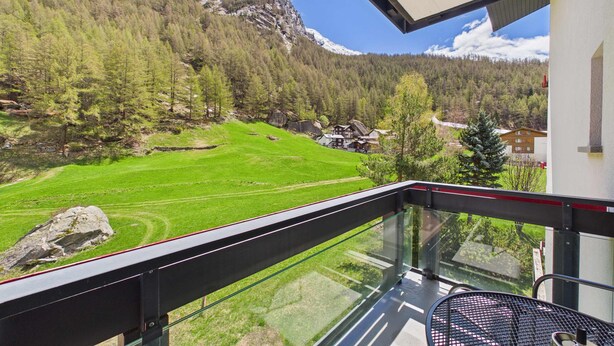 A balcony with a glass railing overlooking a mountainous landscape with green fields and houses. - Alpine Suite near Saas-Fee | Mountain Views (Saas-Almagell)