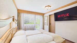 A hotel room with a bed, a television displaying 'NETFLIX,' a window with curtains, and a view of greenery outside. - Alpine Suite near Saas-Fee | Mountain Views (Saas-Almagell)
