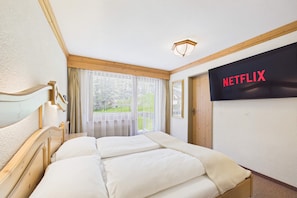 A hotel room with a bed, a television displaying 'NETFLIX,' a window with curtains, and a view of greenery outside.