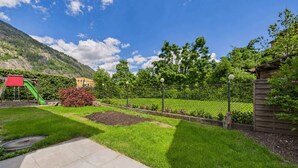 Un jardín con parque infantil, un sendero y montañas al fondo.