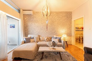 Ein Wohnzimmer mit einer Couch, einem Glastisch, einer Chandelieber und einem Essbereich, der durch eine geöffnete Tür sichtbar ist.