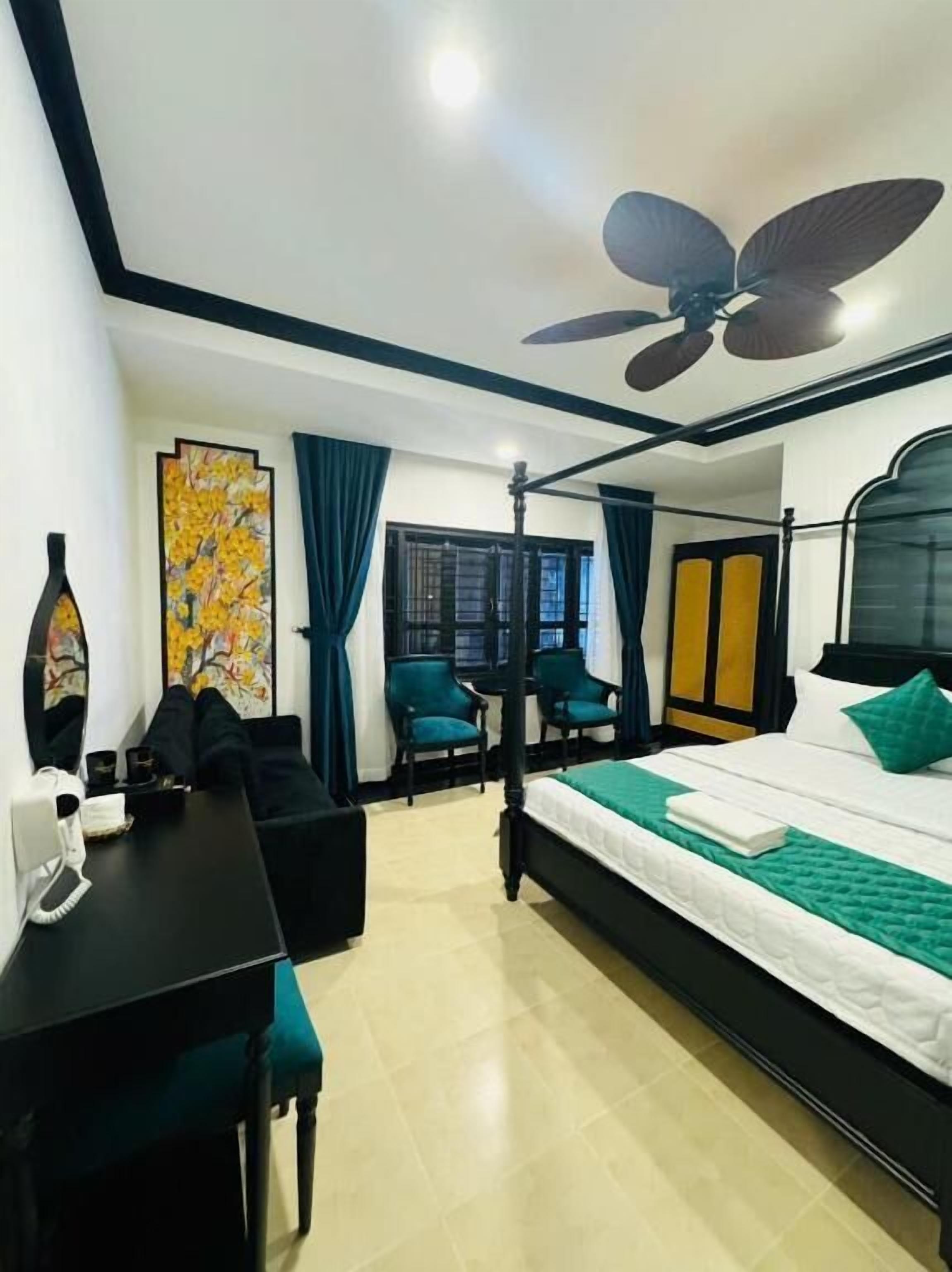 Quarto de hotel com cama, escrivaninha, cadeiras, ventilador de teto, obras de arte, cortinas e uma janela.