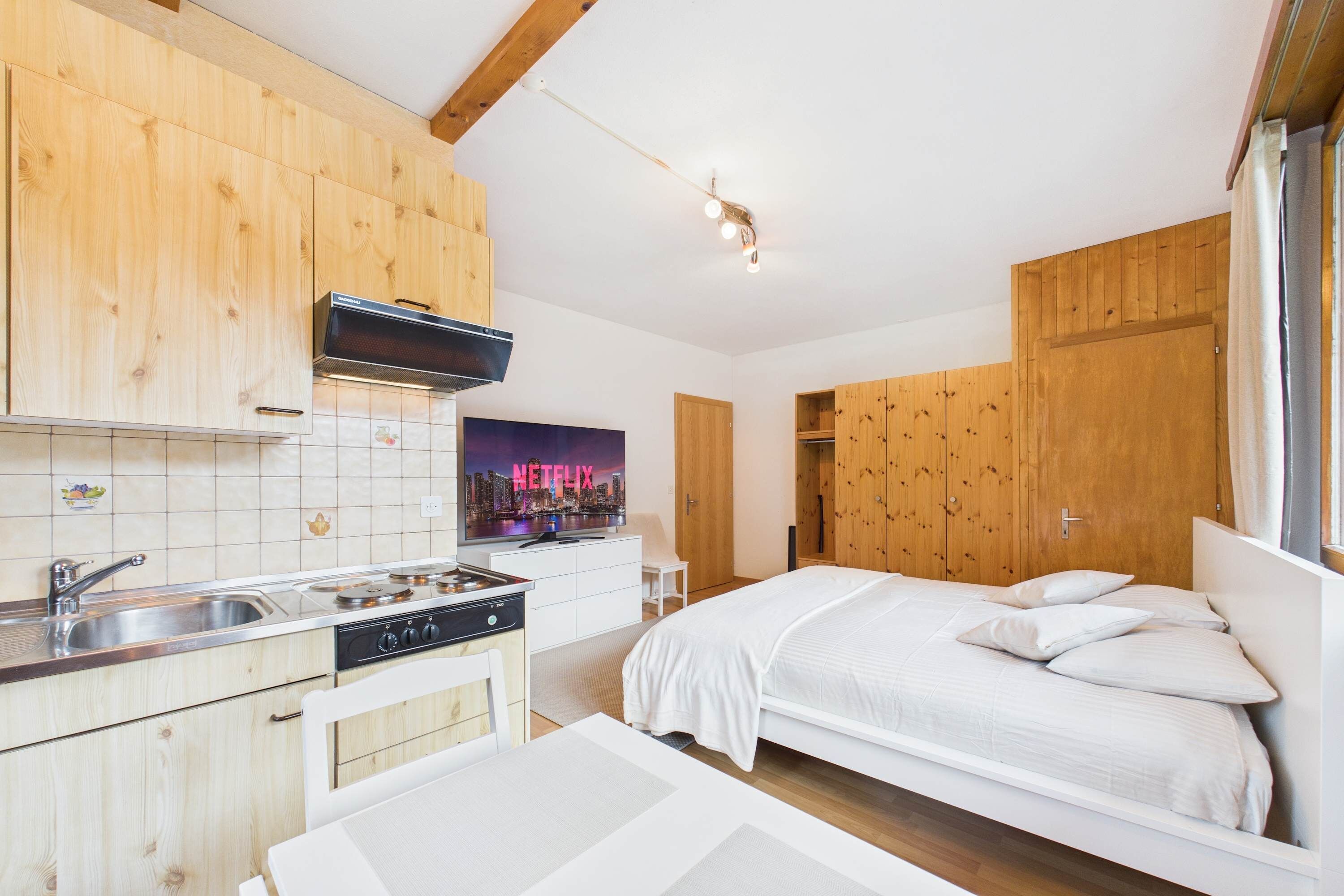Een moderne keuken met houten kastjes, een ingebouwde magnetron en een spoelbak. Er is een bed met wit beddengoed en een poster van een stadsgezicht.