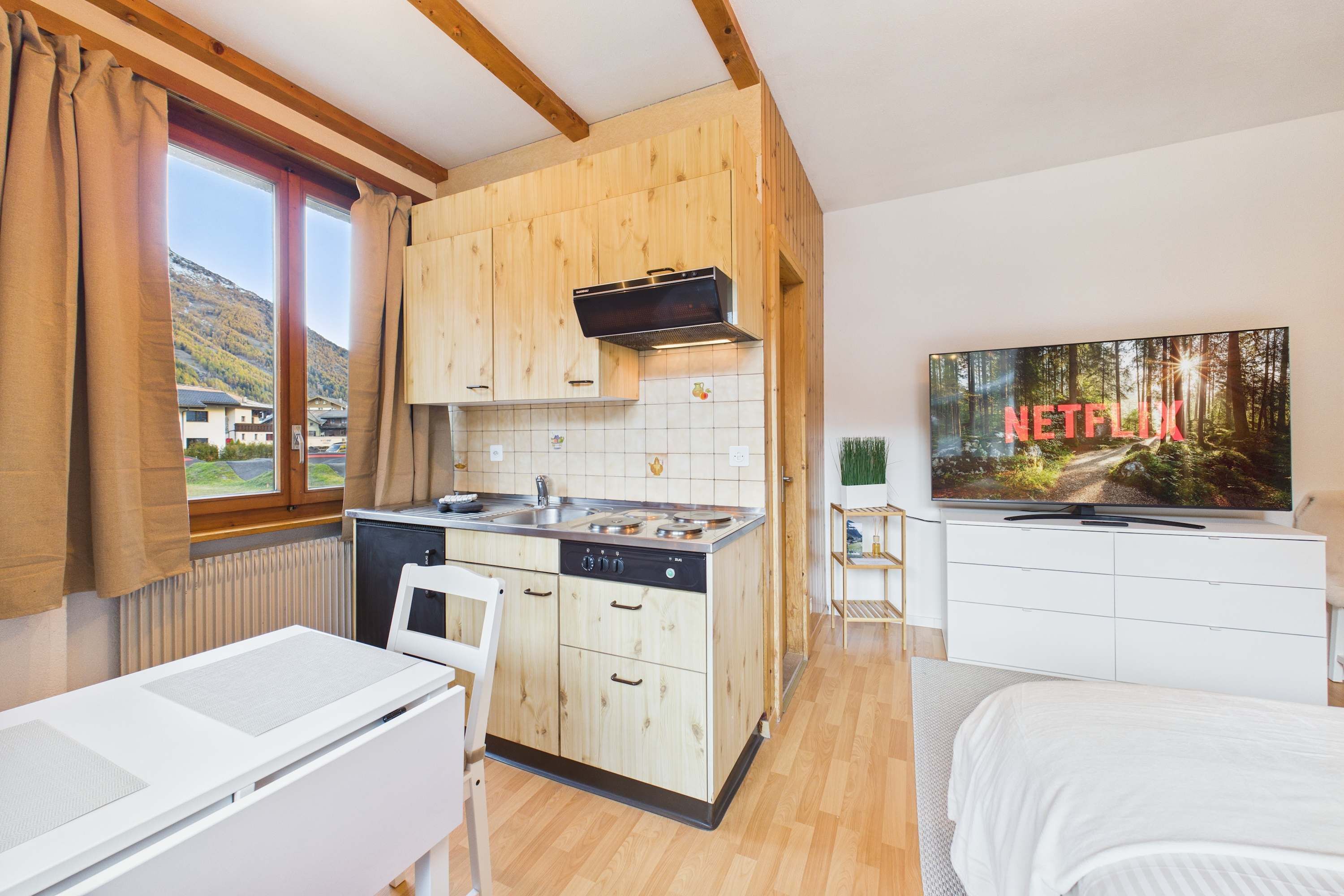 A compact kitchen with wooden cabinets, a stove, and a dishwasher. There's a dining table with chairs, a TV on a stand, and a window with curtains.
