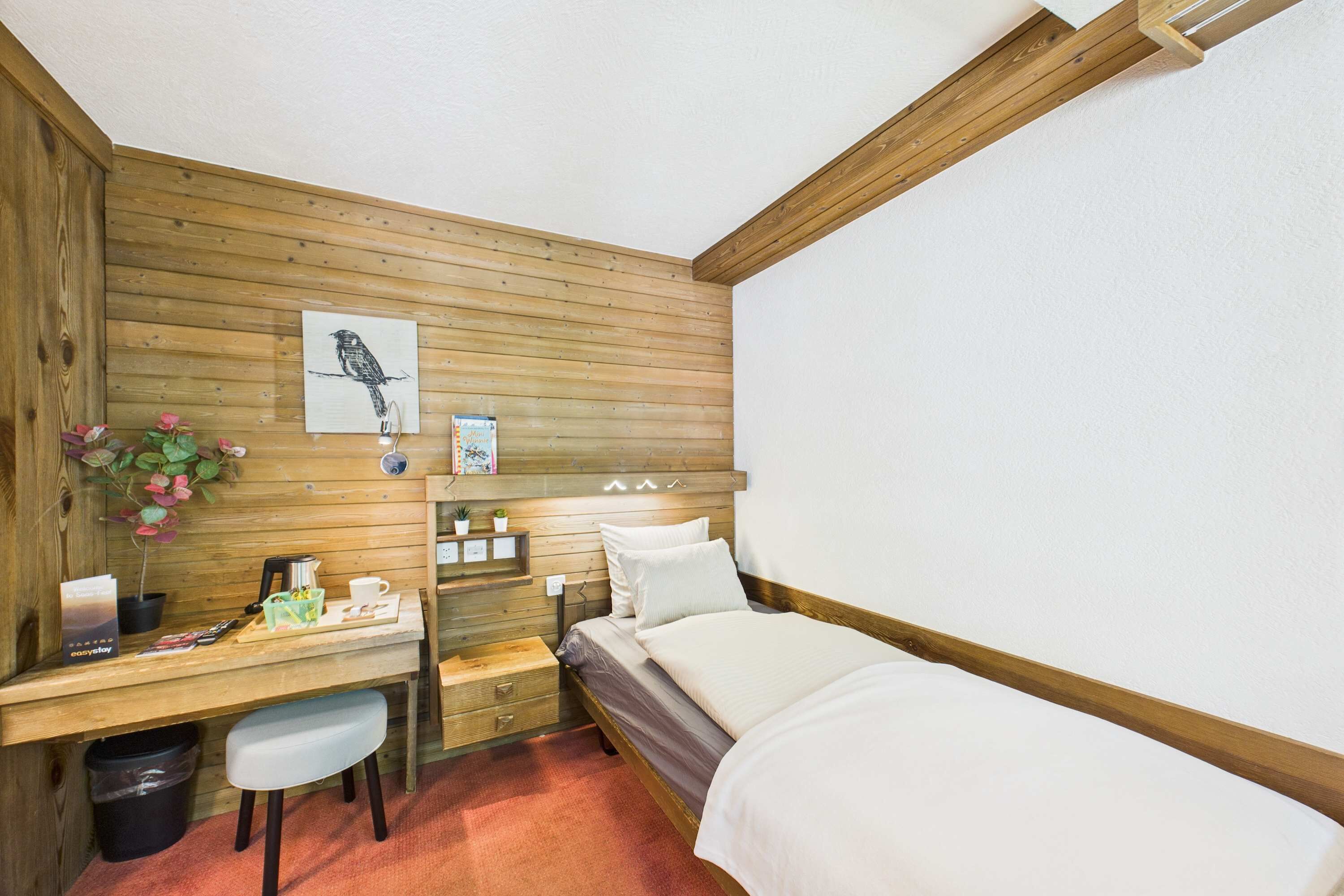 Une pièce avec un mur lambrissé en bois, un lit, un bureau avec une chaise, une poubelle et une petite plante en pot.