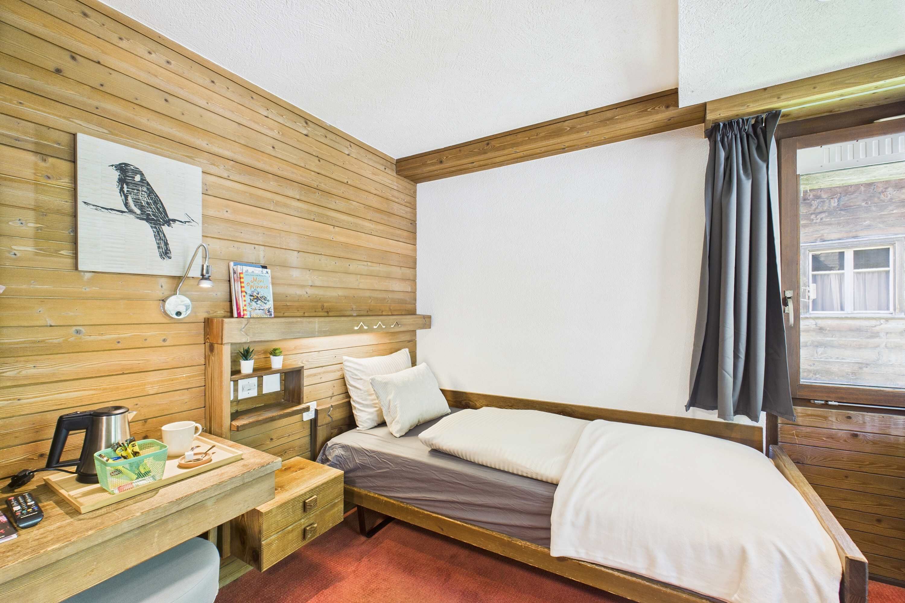 Une pièce aux murs lambrés, comprenant un lit, un bureau, une chaise, une fenêtre avec des rideaux et un tableau accroché au mur.