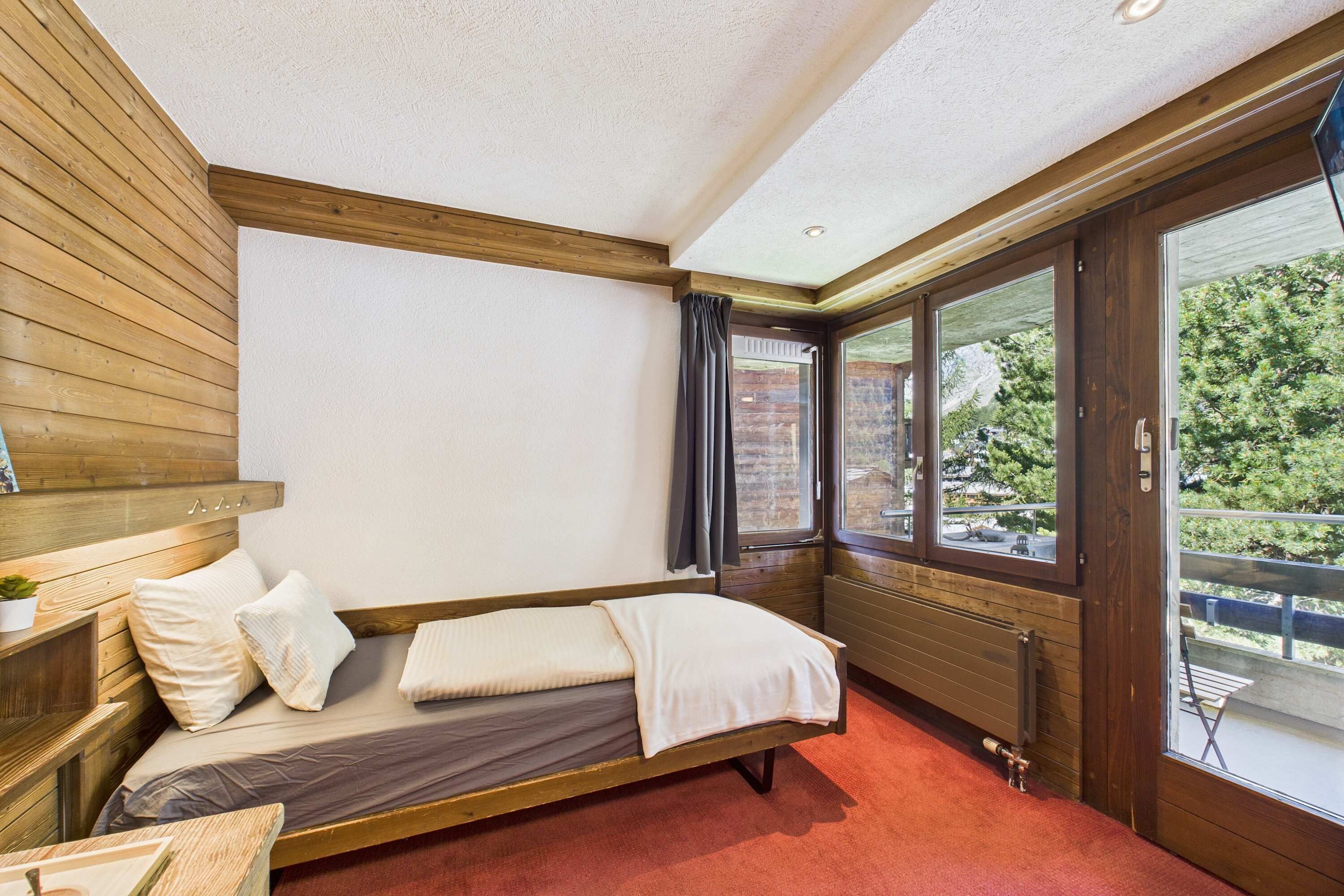A room with a wooden bed, a window with curtains, and a view of greenery outside.