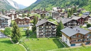 Uma área residencial com vários edifícios de dois andares, alguns com varandas, cercada por vegetação e montanhas ao fundo.