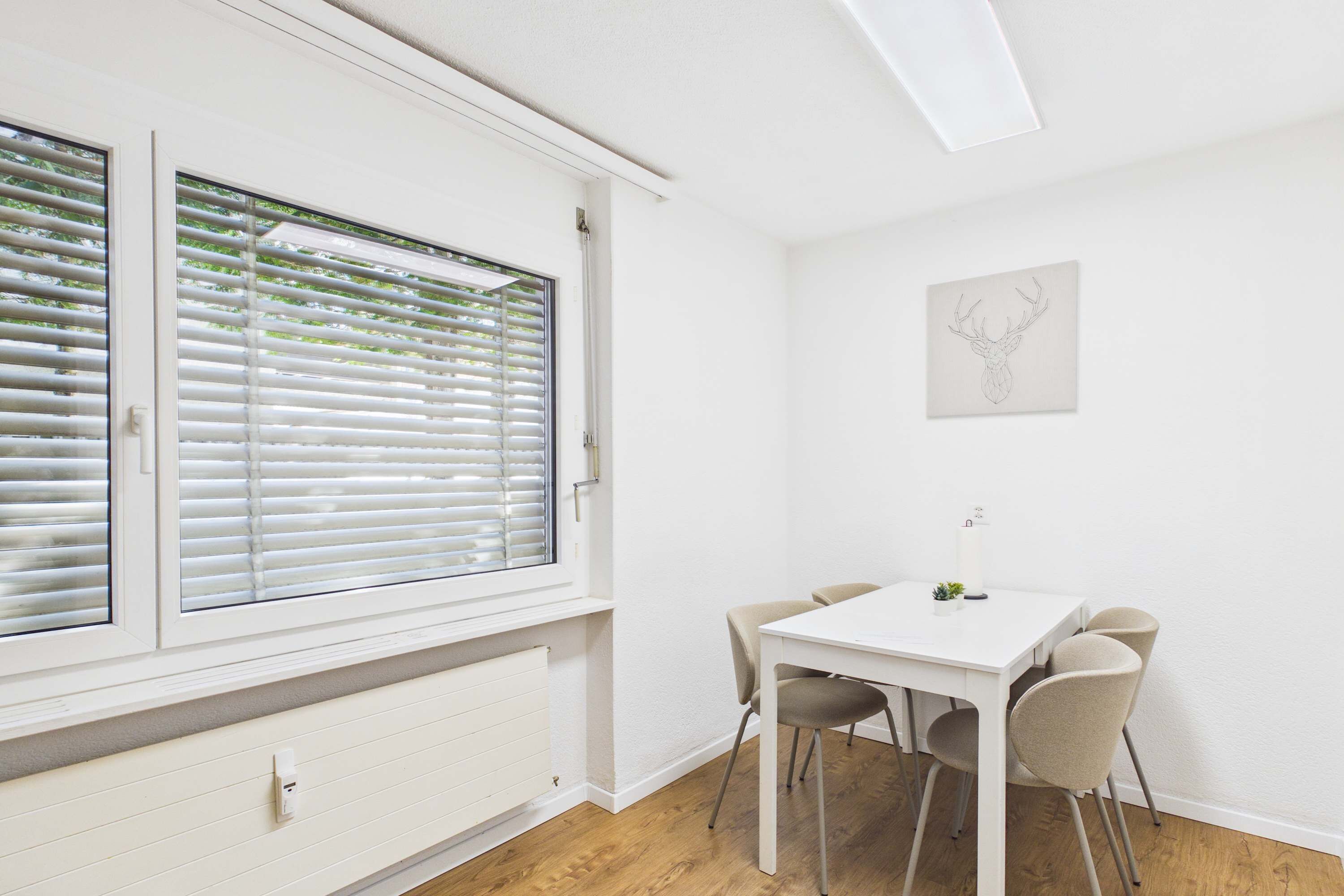 Ein Zimmer mit weißem Tisch und Stühlen, einem Fenster mit Jalousien und einem Wandbild.