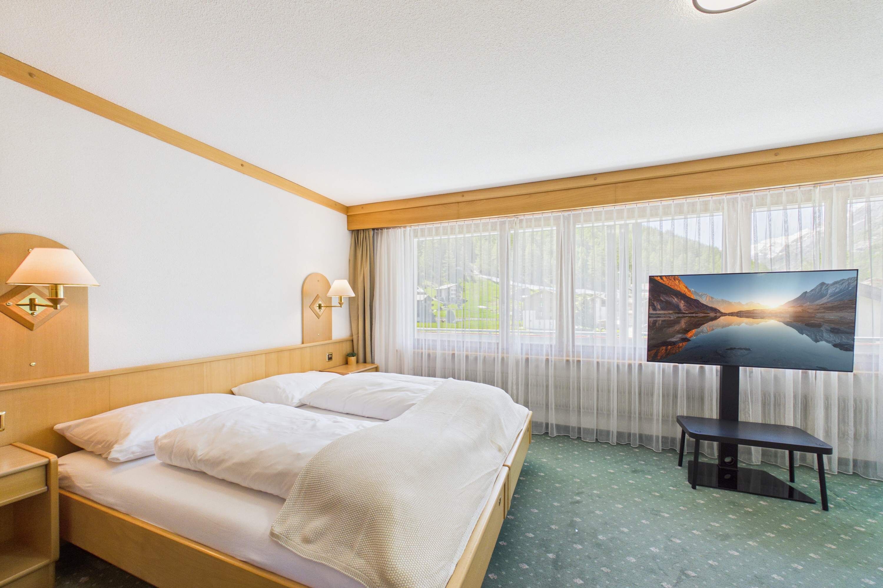 Hotellihuone, jossa on sänky, kaksi yöpöytälamppua, taulutelevisio ja näkymä vuoristomaisemaan ohuen verhon läpi.