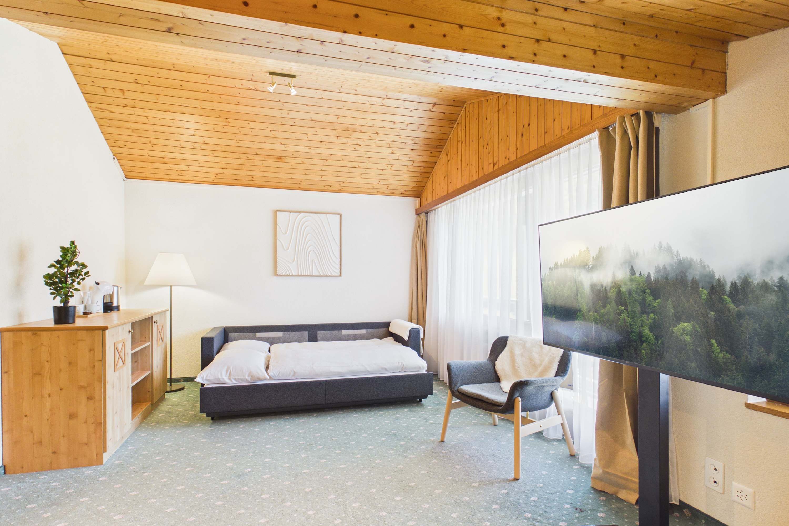 Ein Schlafzimmer mit Bett, Sessel, Schreibtisch, Fernseher und einem Fenster mit Vorhängen.