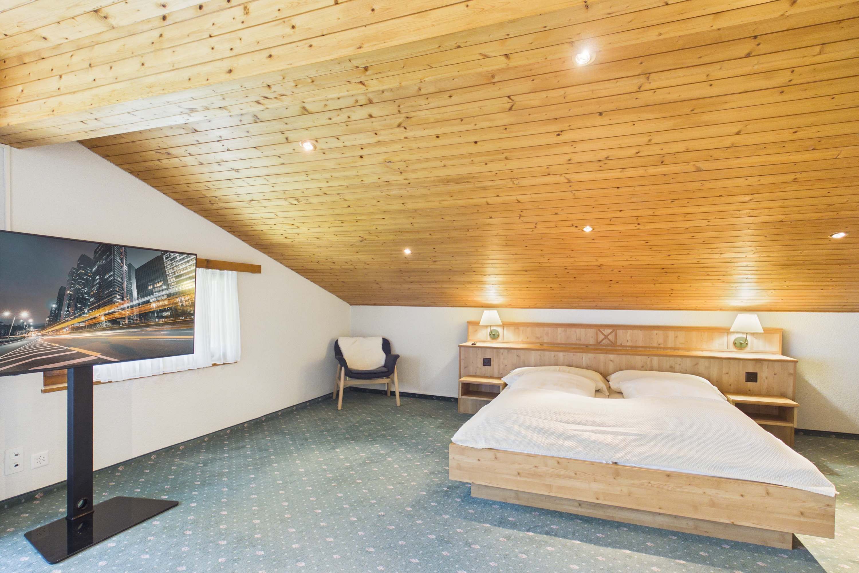 A bedroom with a wooden ceiling, a bed, a desk, a chair, and a window with a cityscape view.