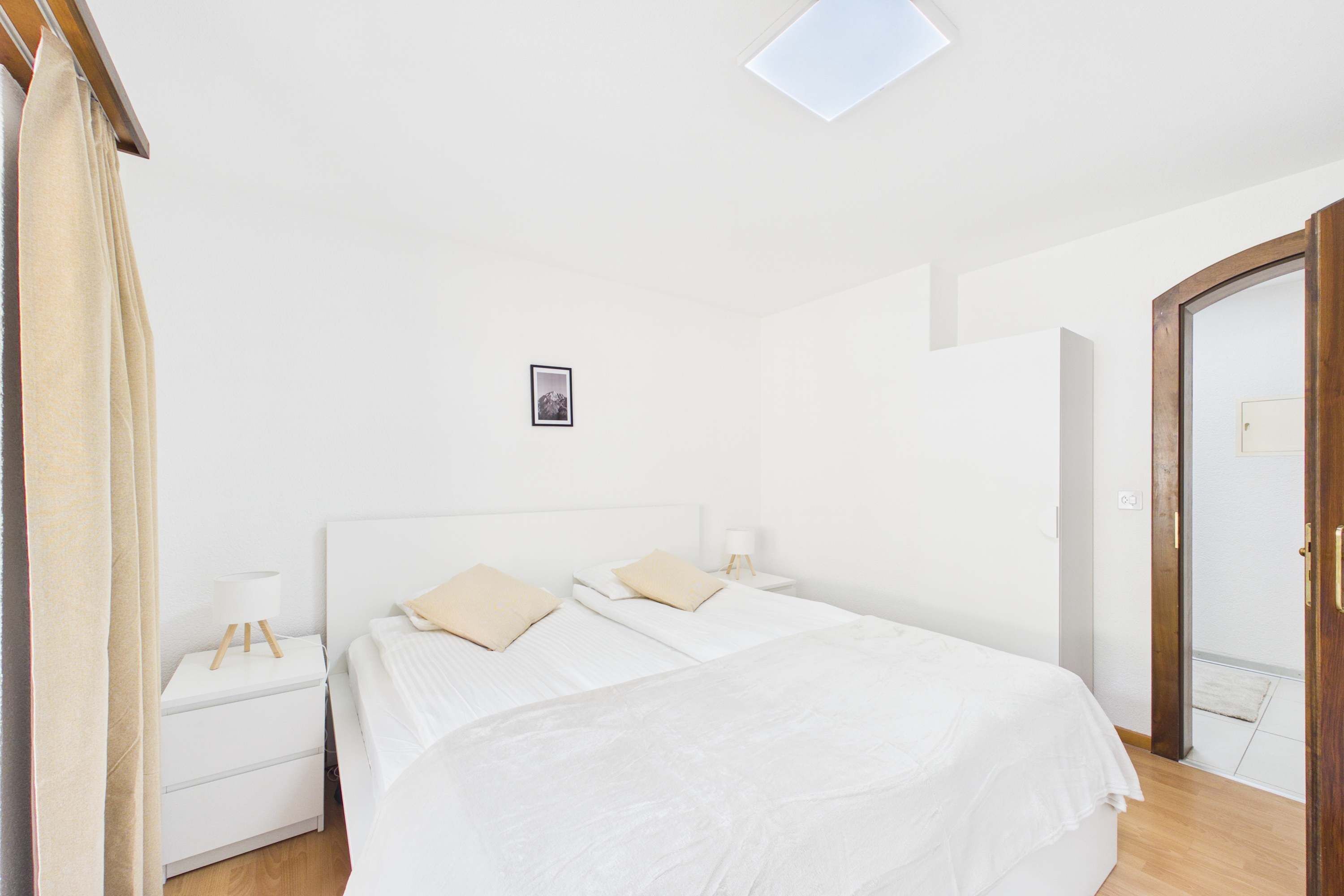 Un dormitorio con cama, mesitas de noche, una lámpara de noche, un cuadro en la pared y una puerta.