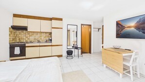 A hotel room with a bed, a desk, a chair, a small table, a mirror, and a kitchen area. - Charming Studio with Mountain Views in Brig (Gamsen)
