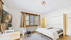 Un dormitorio con cama, escritorio, silla, radiador, ventana con cortinas y un cuadro en la pared.