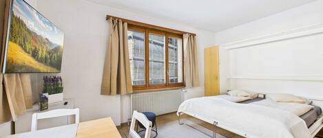 Ein Schlafzimmer mit Bett, Schreibtisch, Stuhl, einem Fenster mit Vorhängen, einer Heizung und einem Bild an der Wand.