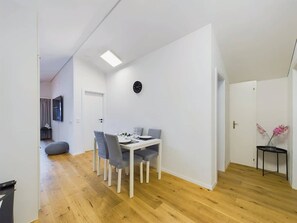Een eethoek met een witte tafel, grijze stoelen, een klok aan de muur, en een bijzettafeltje met bloemen.