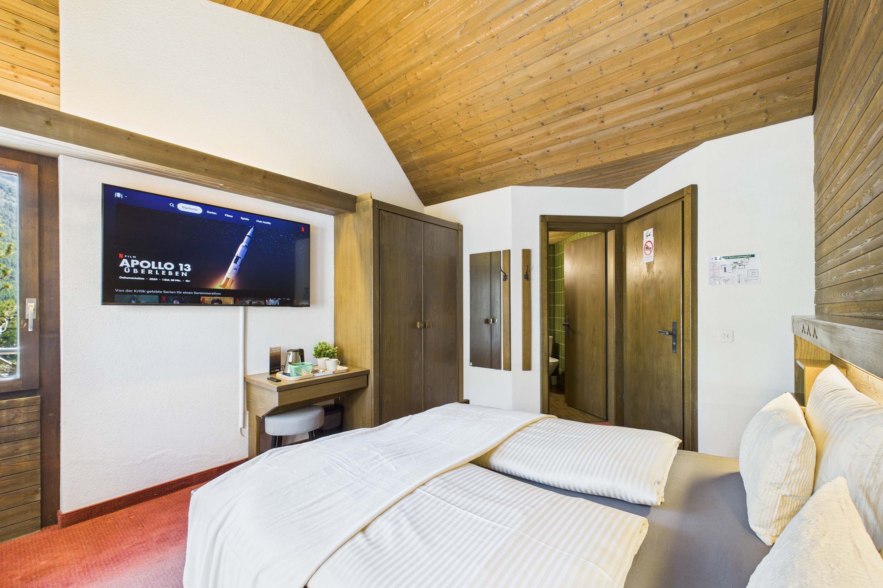 Habitación de hotel con televisión montada en la pared, un escritorio con silla, una cama con dos almohadas y techo de madera.