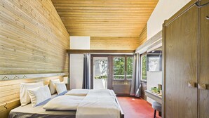Un dormitorio con techo de madera, una cama con ropa blanca, un armario de madera y vistas a través de un ventanal.