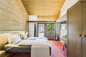 Un dormitorio con techo de madera, una cama con ropa blanca, un armario de madera y vistas a través de un ventanal.