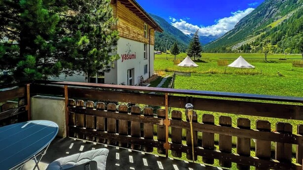 A balcony with a table and chairs, a view of a mountain landscape, and a building with 'yasumi' signage. - Mountain View Apartment Steps from Zermatt (Täsch)