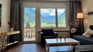Una habitación con sofá, una silla, una mesita de centro, una lámpara y vistas a montañas a través de ventanas amplias.