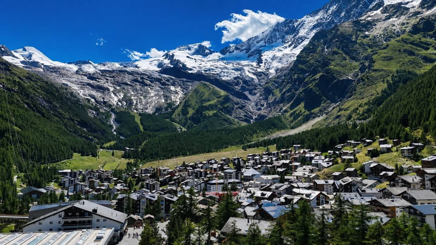 A mountain village with buildings clustered at the base, surrounded by snow-capped peaks and dense forests.