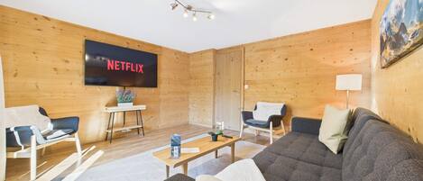 A cozy living room with a large flat-screen TV displaying 'NETFLIX,' a sofa, two armchairs, a coffee table, and a small table with a potted plant.