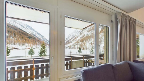 Une pièce offrant une vue sur un paysage montagneux enneigé grâce à de grandes fenêtres, avec une balustrade en bois et un canapé gris.
