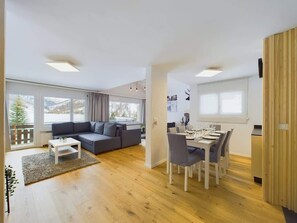 Een moderne woonkamer met een bank, eettafel en stoelen, grote ramen en een houten vloer.