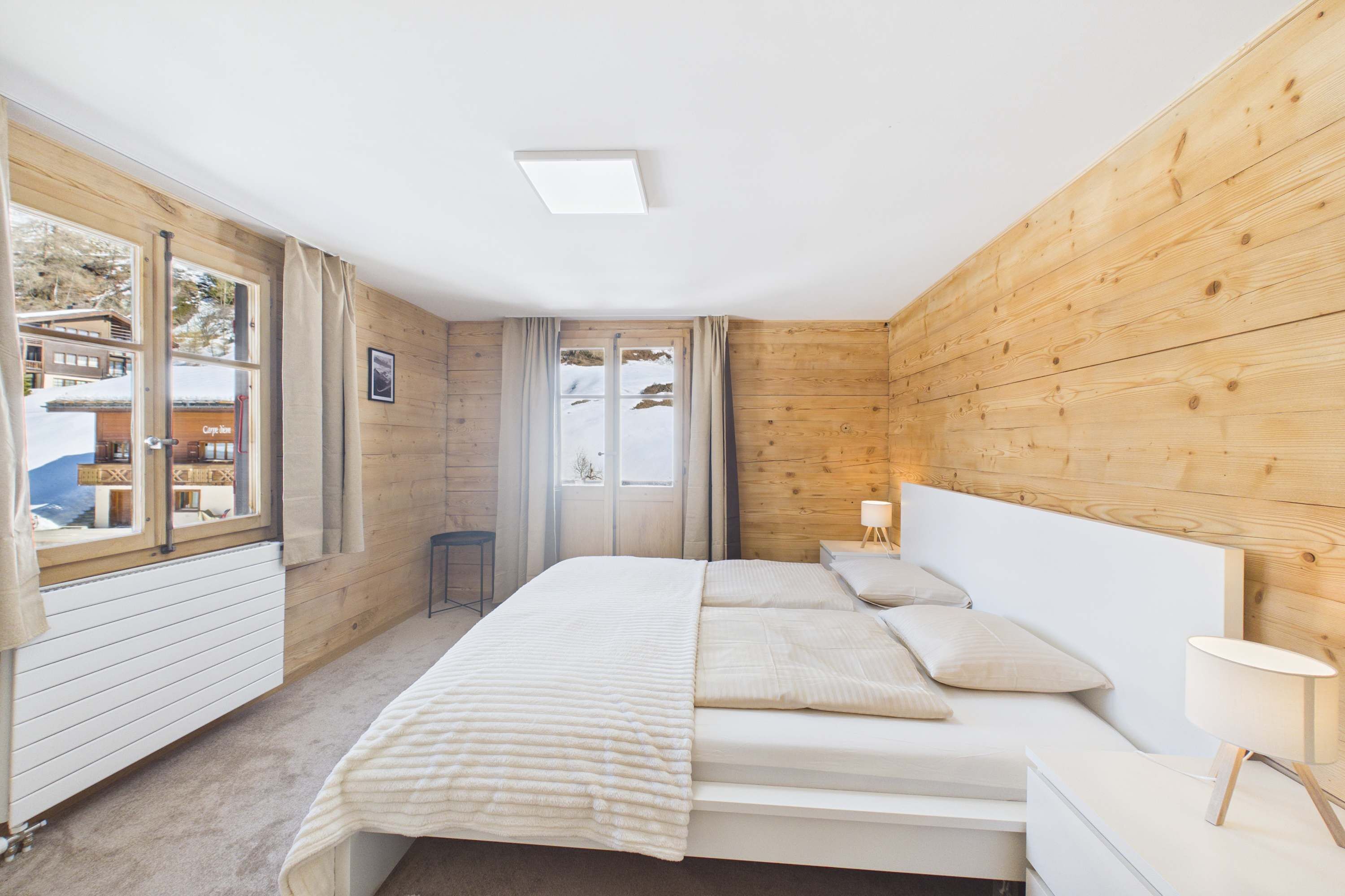 A bedroom with a wooden wall, a bed with white bedding, a bedside table with a lamp, and a window with curtains.