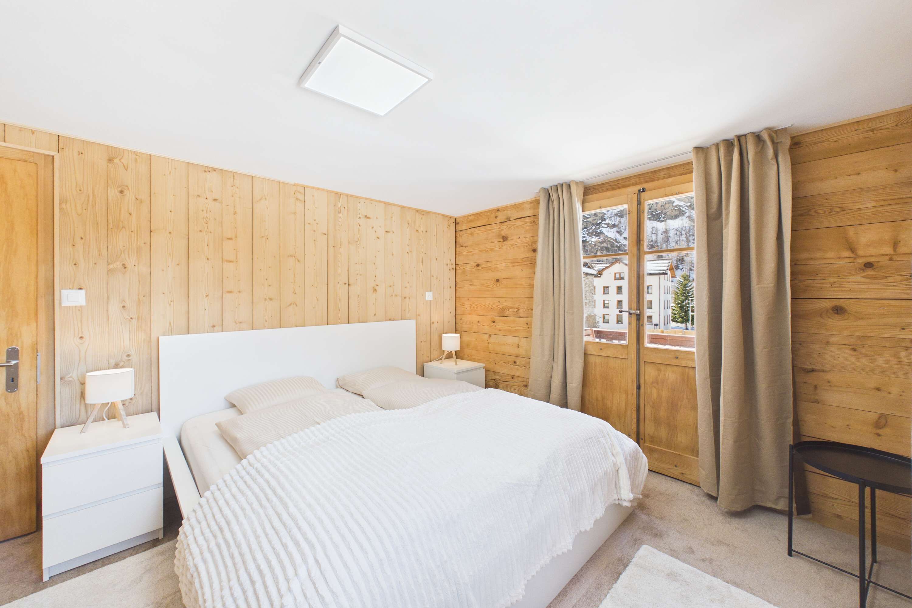 Ein Schlafzimmer mit Holzwänden, einem Bett, zwei Nachttischen mit Lampen, einem Fenster mit Vorhängen und Blick auf Gebäude.