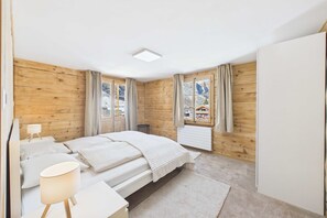 A bedroom with wooden walls, a bed, a bedside table with a lamp, and a view of mountains through the window.