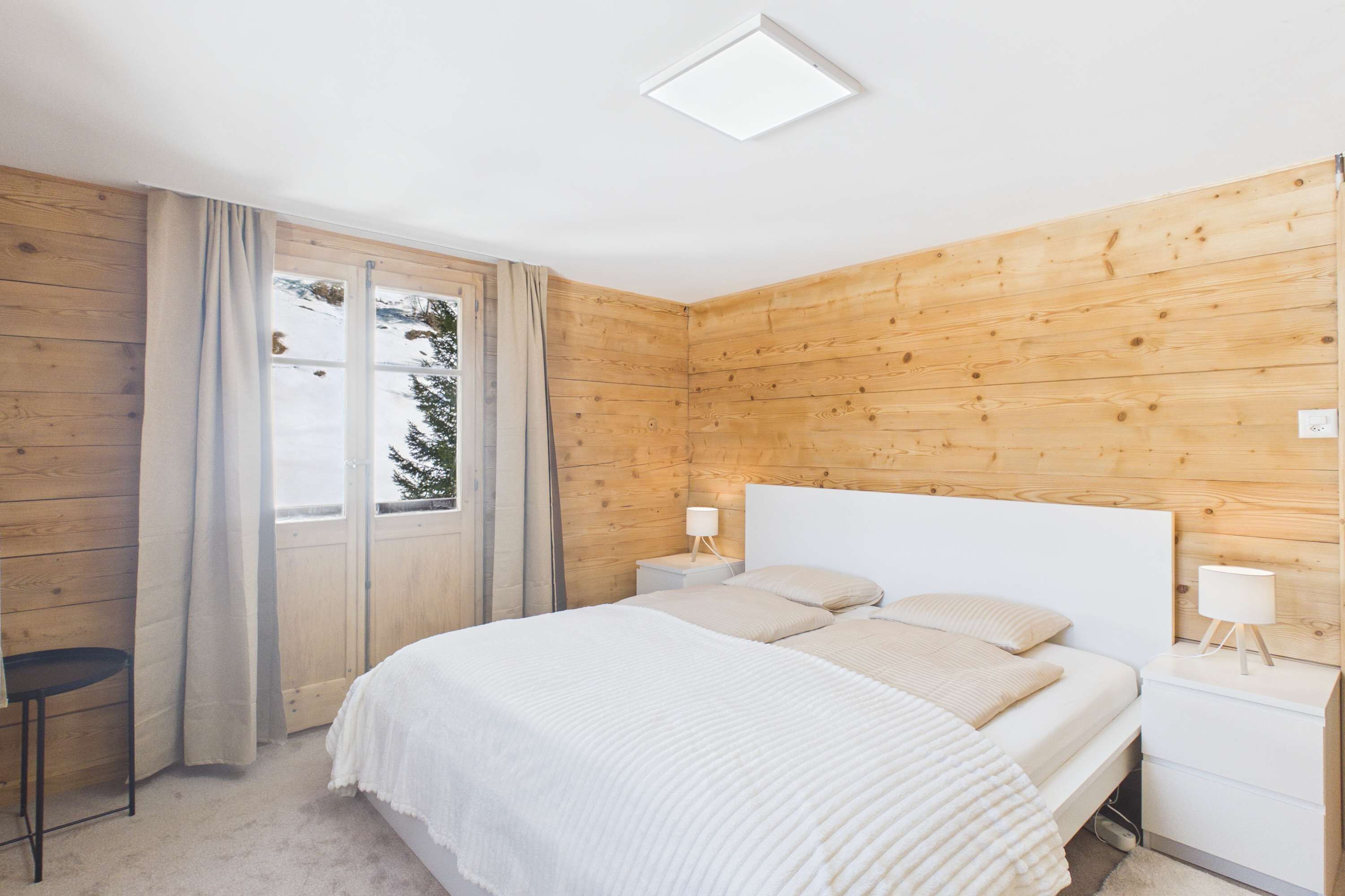 Une chambre avec un mur lambrissé en bois, un lit avec une literie blanche, une table de chevet avec une lampe et une fenêtre avec des rideaux.