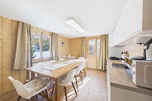 A dining area with a wooden table and chairs, a kitchen with a microwave, and large windows with curtains.