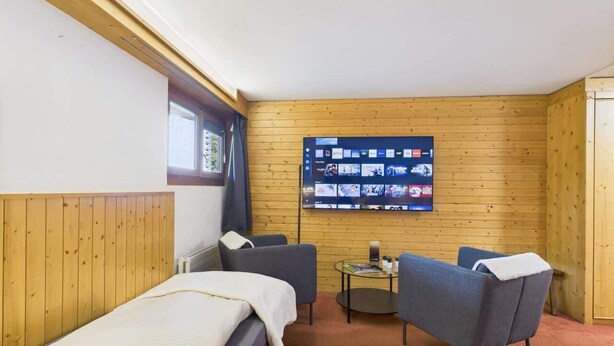 Una habitación con una cama, dos sillones, una mesita y un televisor en la pared que muestra varios canales.