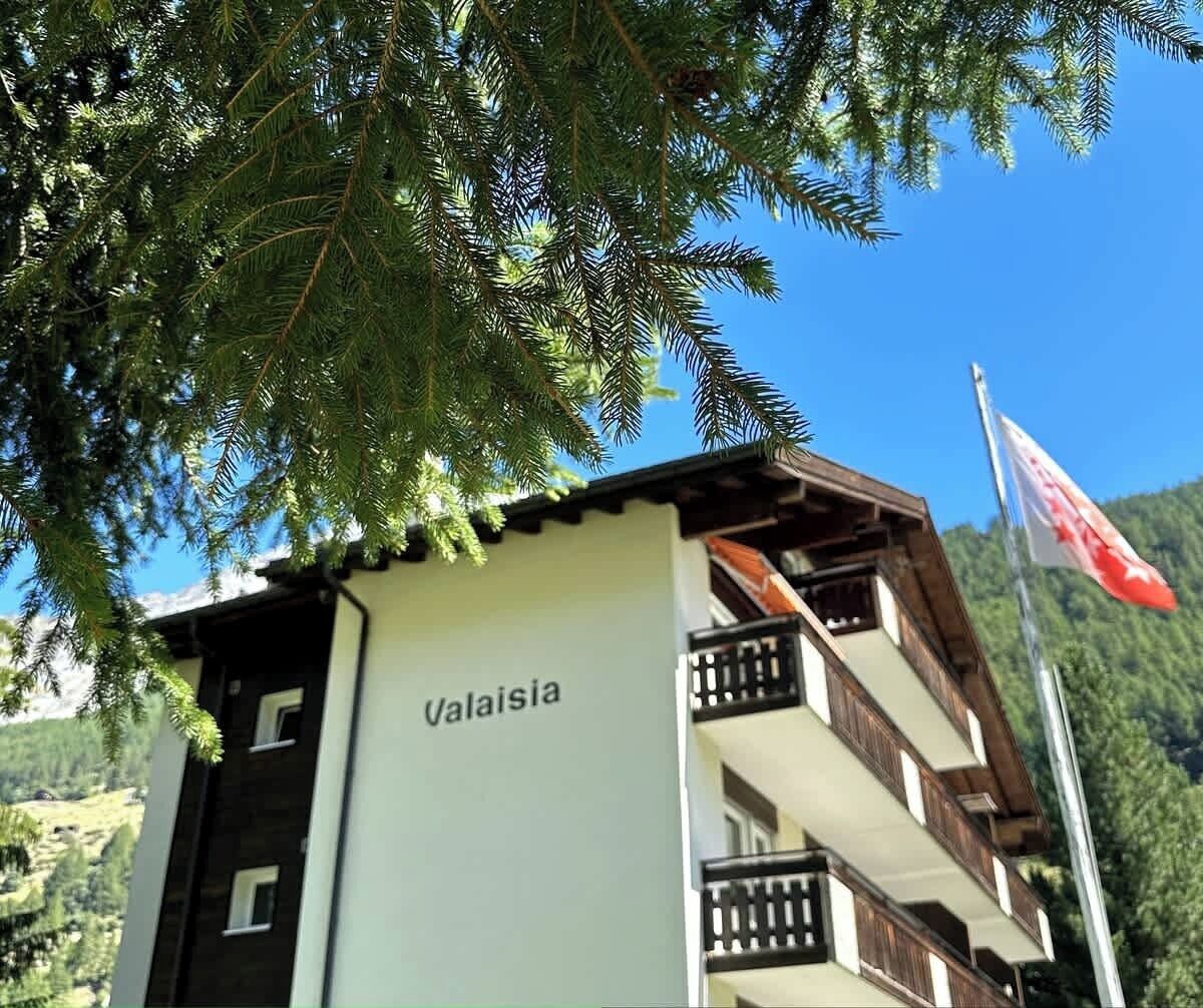 Ein Gebäude mit Balkonen und einer Flagge, beschriftet mit 'Valaisia', vor einer Bergkulisse.