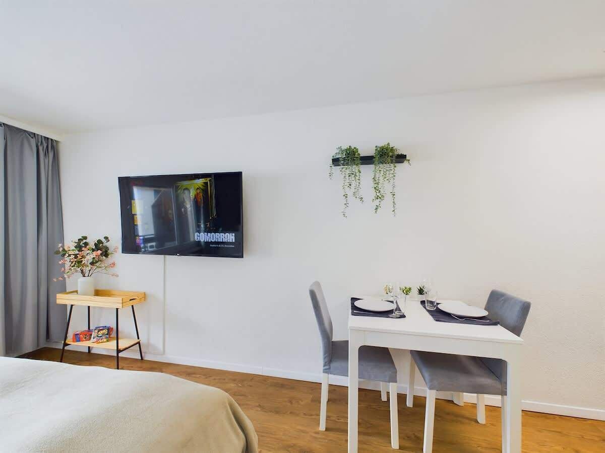 Ein modernes Hotelzimmer mit einem Bett, einem an der Wand montierten Fernseher, einem Esstisch mit Stühlen und einer Pflanze, die von der Decke hängt.