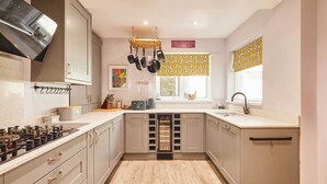 A kitchen with a gas stove, hanging pots, a built-in wine cooler, and a window with patterned curtains. - Host & Stay | The Mews (Rothbury)