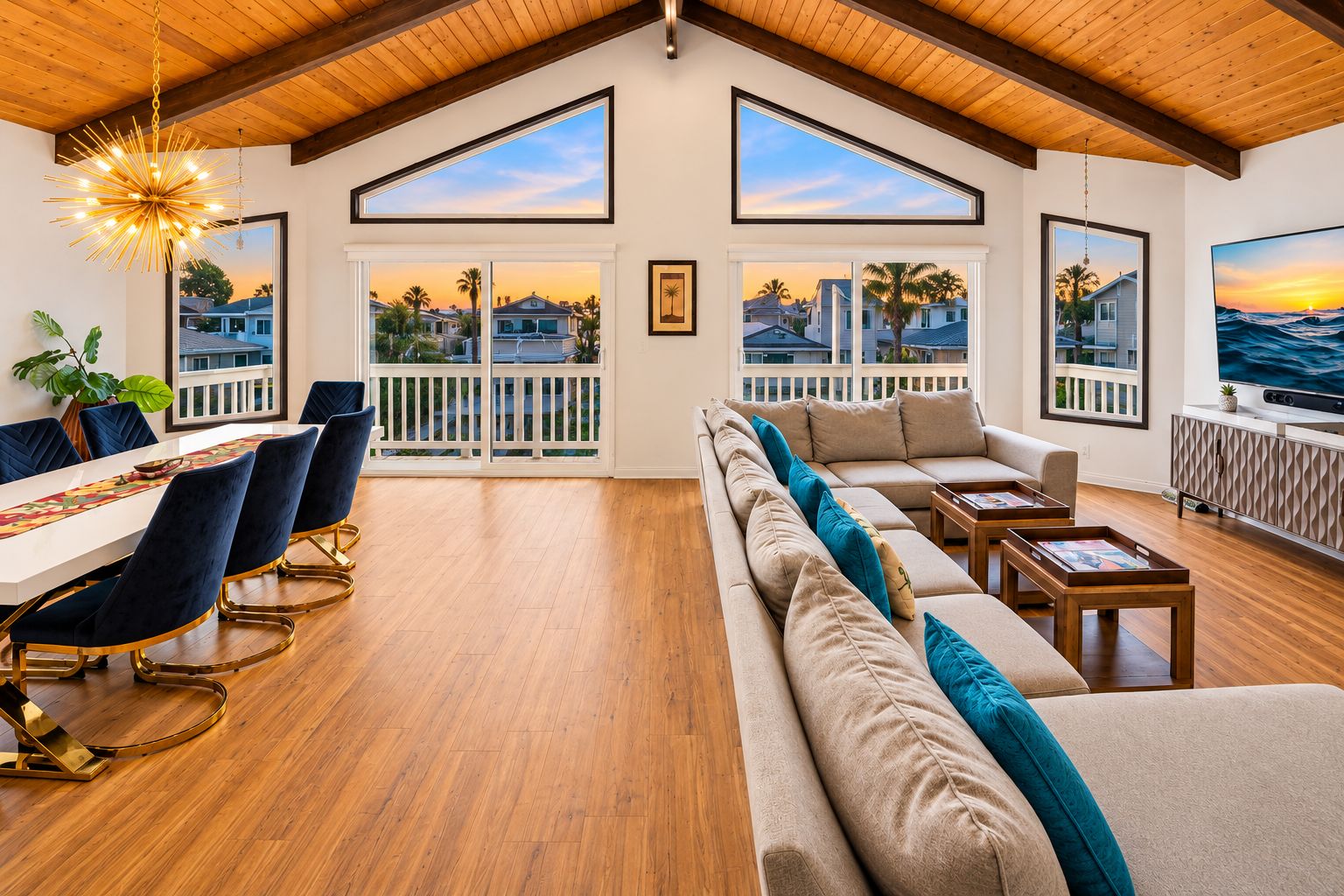 A spacious living area with a high ceiling, wooden beams, a dining table with chairs, a sofa with cushions, and large windows offering views of a residential neighborhood.