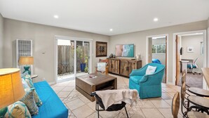 A spacious living room with a blue sofa, a wooden coffee table, and a teal armchair. There is a sliding glass door leading to a patio area with potted plants. - 25% Off Sept 3-6! Mins 2 Beach, Rooftop Patio View (Oxnard)