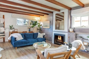 Ein Wohnzimmer mit Kamin, einem blauen Sofa, Holzbalken und Blick ins Freie.