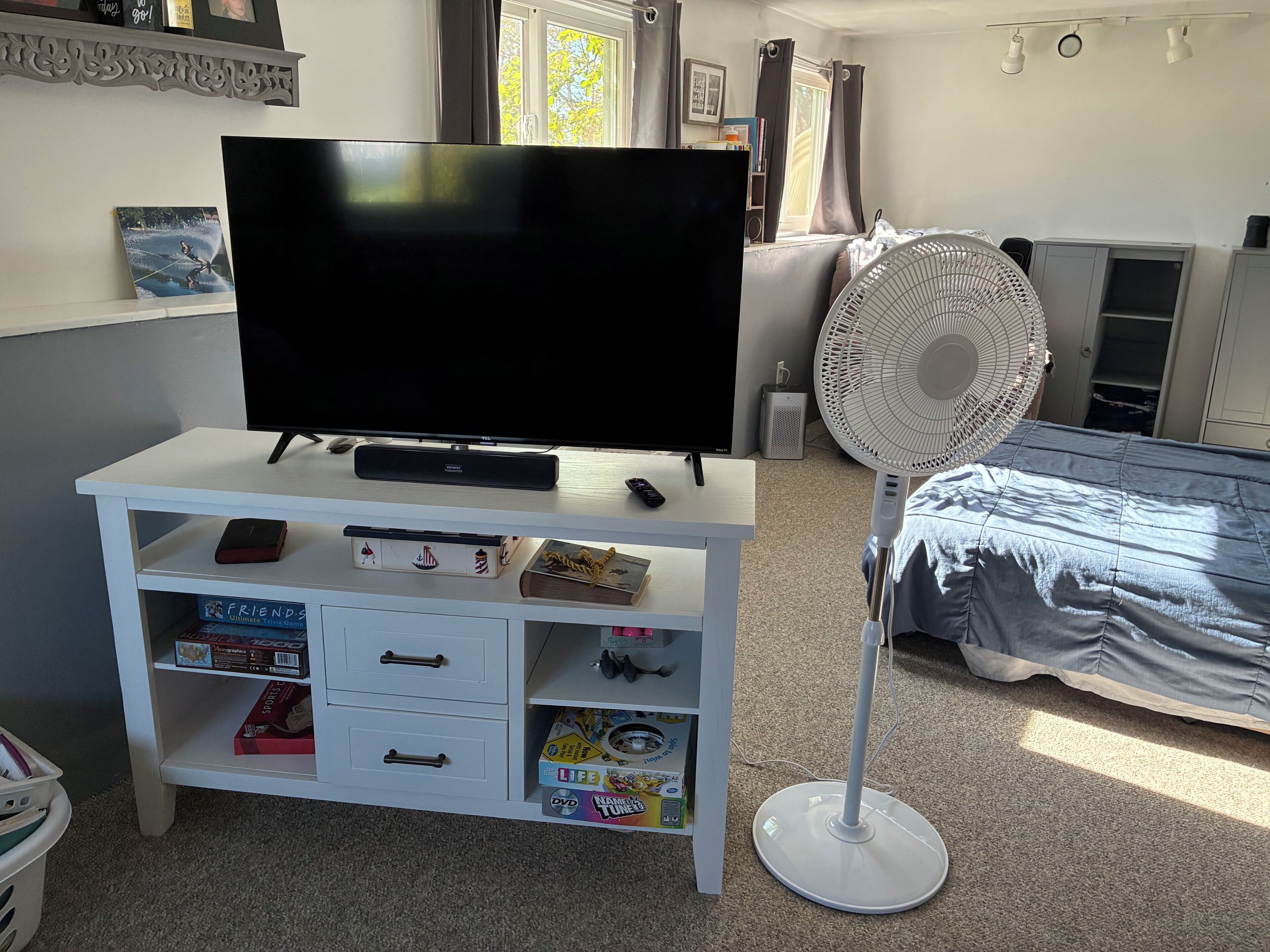 A bedroom with a TV on a white stand, a fan, a bed with a blue cover, and a shelf with various items.