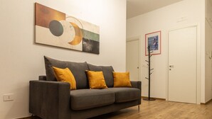 Un sofá gris con tres cojines color mostaza, suelo de madera, un perchero y una puerta en una habitación con pared blanca.